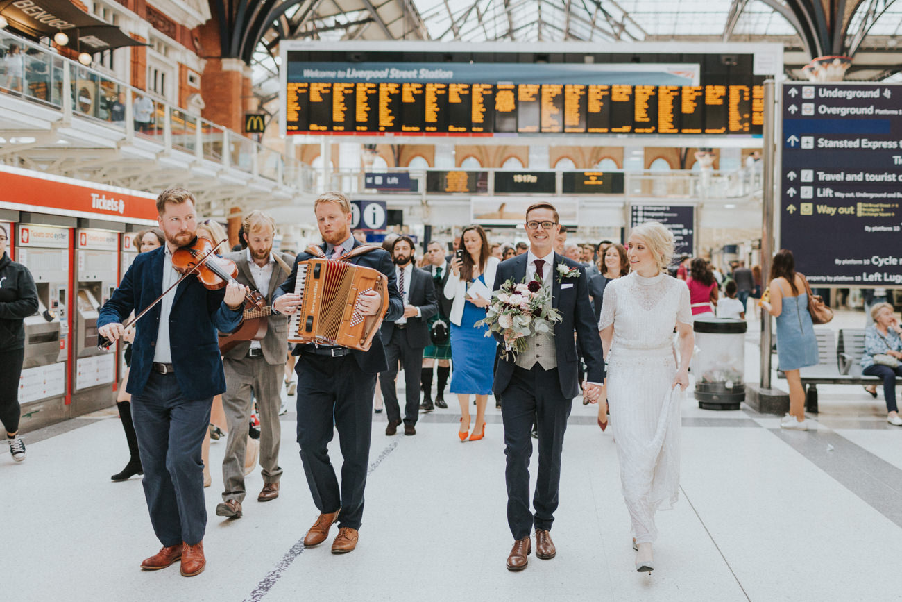 Bride and groom walking with their wedding party and musicians through Liverpool St Station in London.